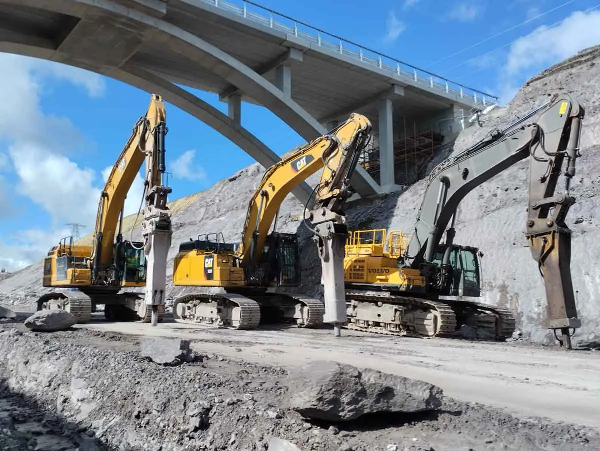 Three large excavators with hydraulic breakers parked under a concrete bridge arch